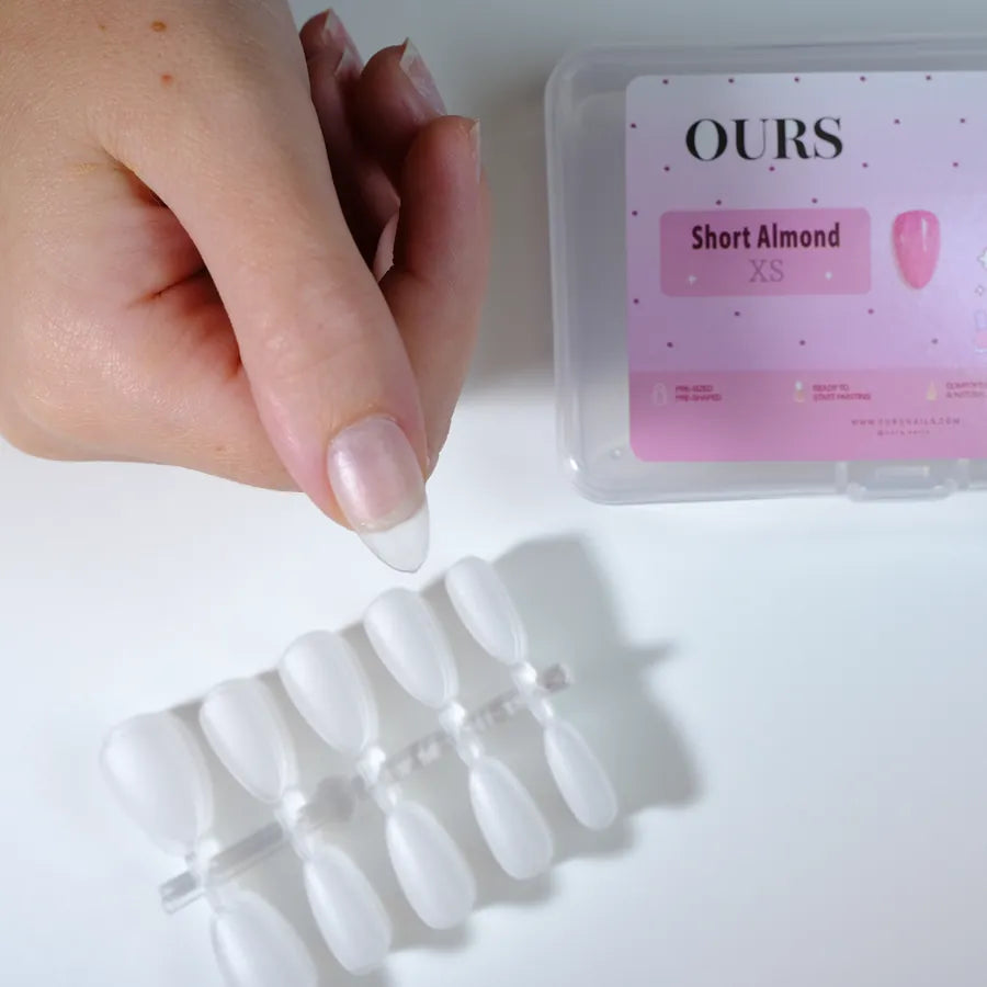 Holding a clear short almond nail tip above a tray, OURS box behind